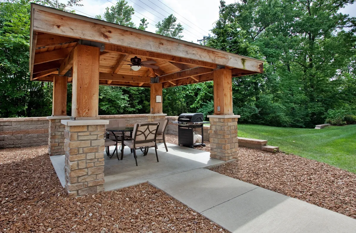 A spacious outdoor area featuring a wooden gazebo with sturdy stone pillars, a grill, and an outdoor dining set. The scene is set for a relaxed gathering surrounded by greenery, ideal for family barbecues or casual meals outside.
