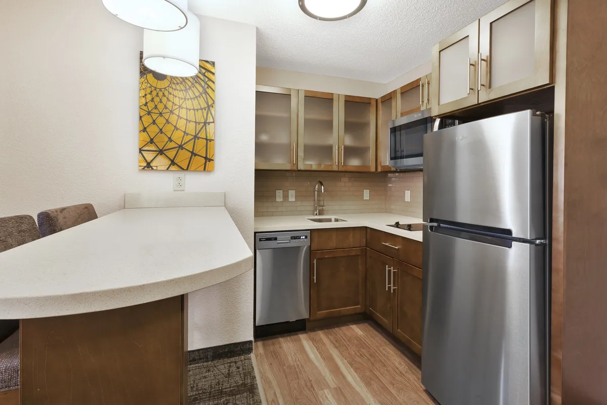 This image shows a modern kitchen with a sleek countertop equipped with bar seating, stainless steel appliances such as a refrigerator, stove, and dishwasher, and decorative artwork on the wall. The space is designed for both cooking and socializing, with a welcoming and contemporary atmosphere that suggests it's ideal for hosting gatherings or enjoying family meals.