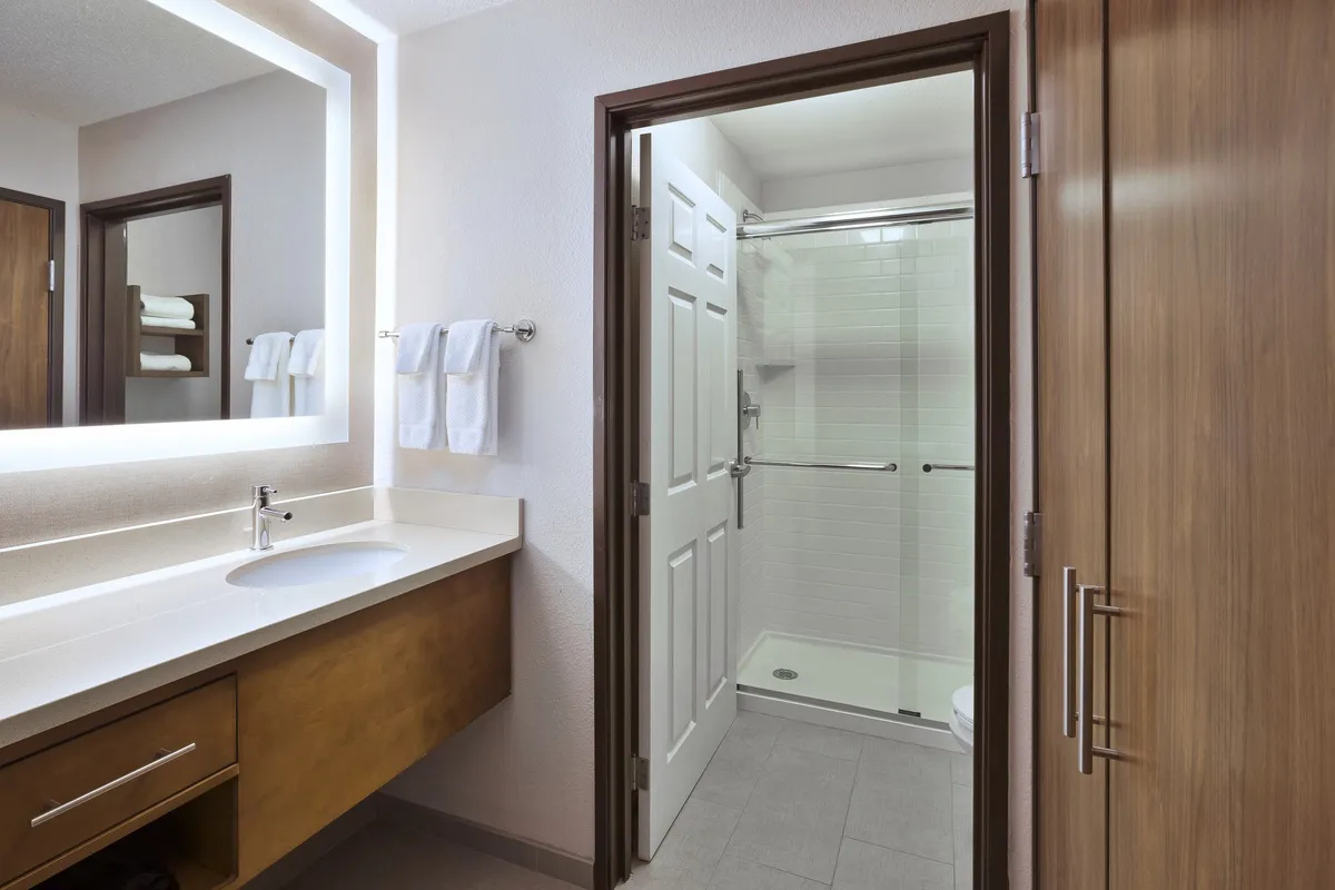 A modern, sleek bathroom with a well-lit vanity, sink, large mirror, shower area, and neatly arranged fresh towels. The serene mood and spotless fixtures suggest a premium experience with thoughtful amenities.