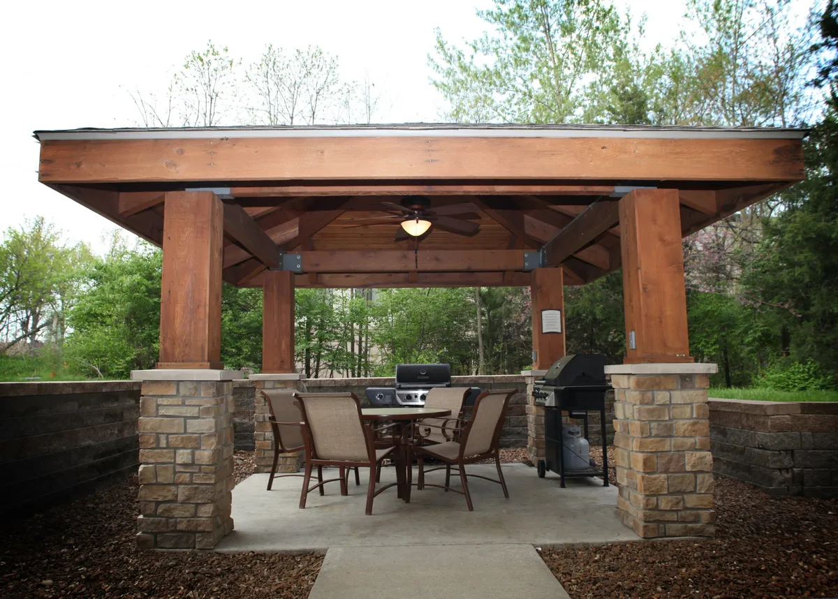 An exterior view of a wooden pavilion equipped with an outdoor dining table and a grill setup. The space is designed for group meals and casual get-togethers, surrounded by natural elements that make it ideal for spending time outside with loved ones.