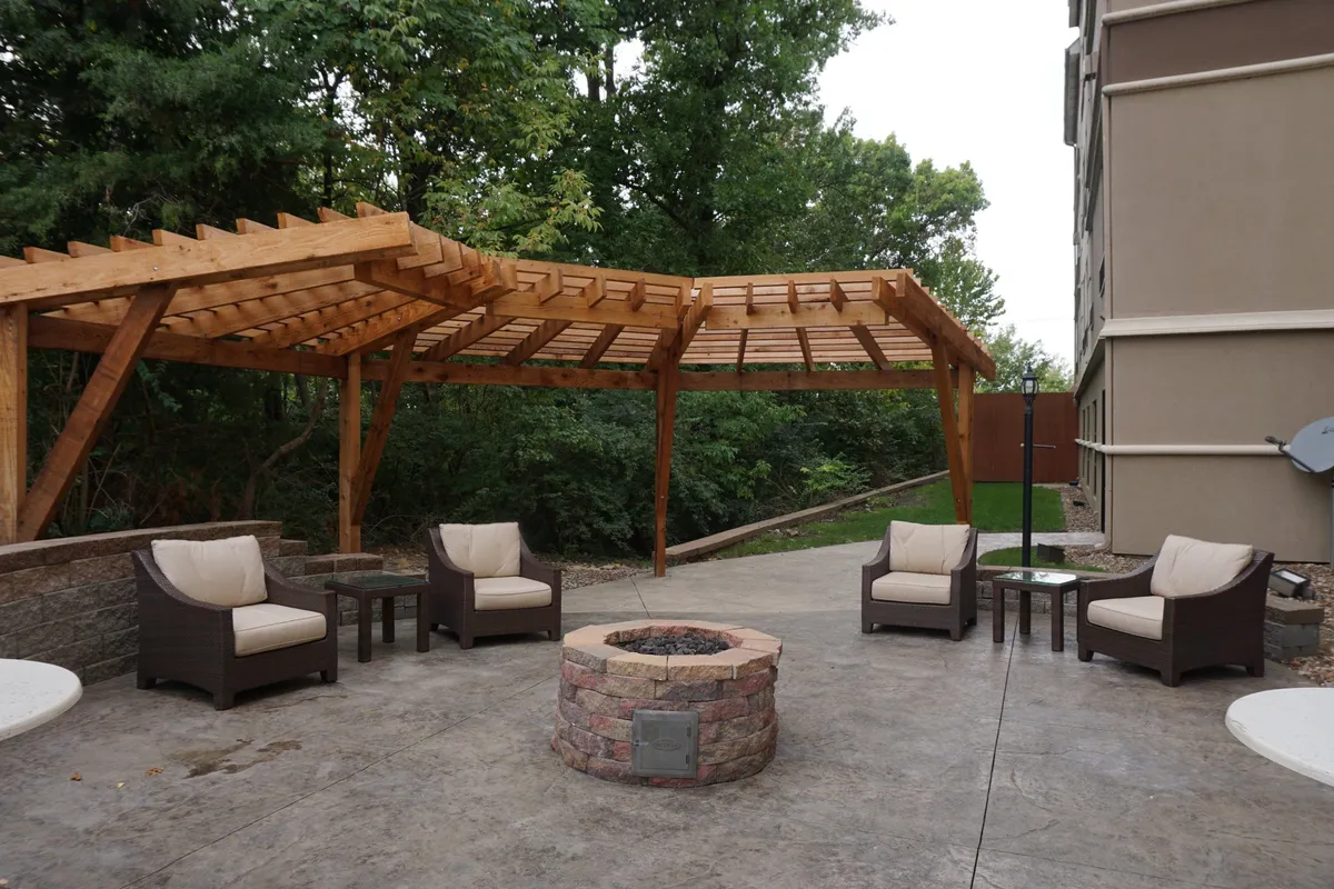 A cozy outdoor scene with a pergola sheltering a fire pit, surrounded by comfortable seating and lush greenery. The setting evokes evenings spent sharing stories, making s'mores, or simply unwinding together in a warm, communal atmosphere.