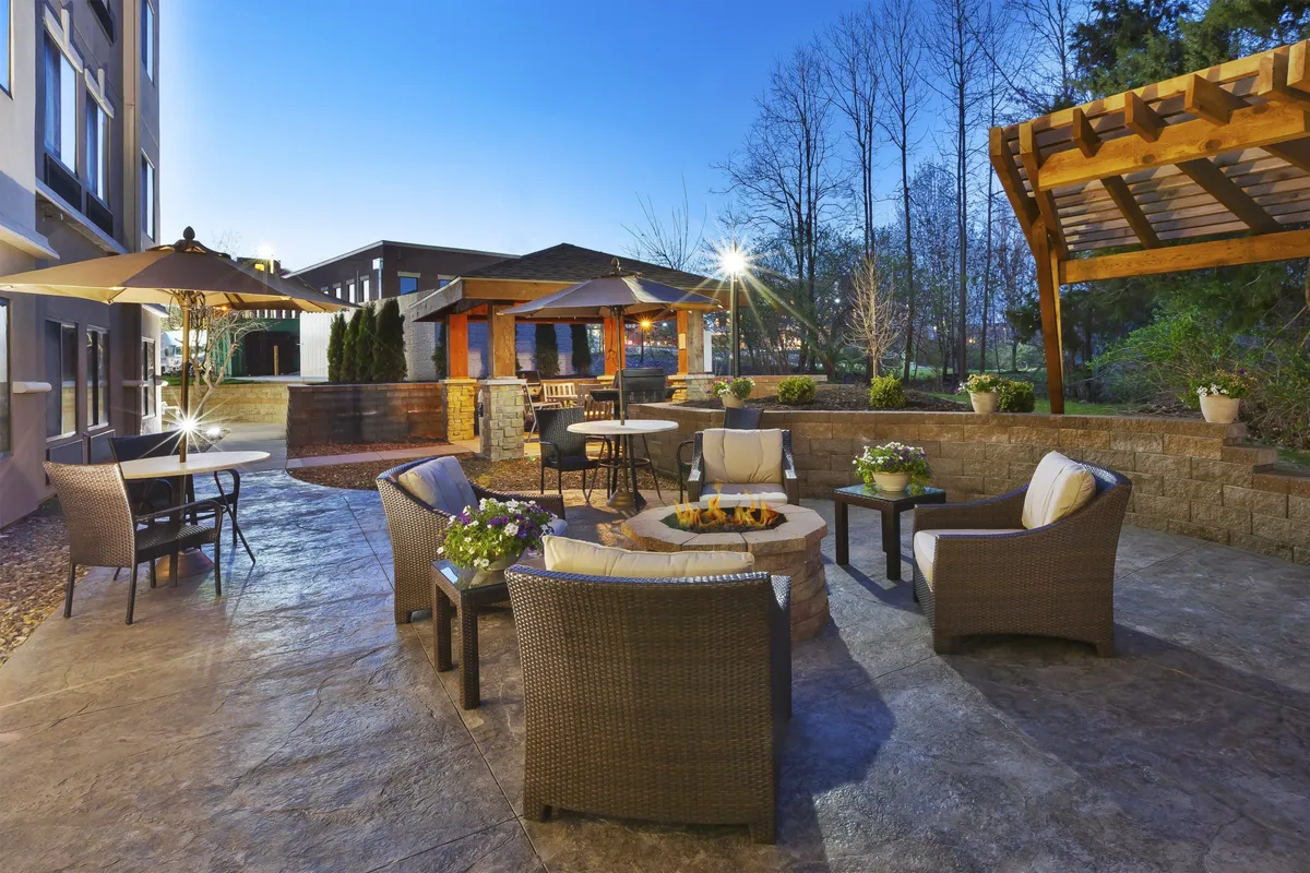 An exterior evening scene highlights a cozy outdoor seating area around a fire pit, under a pergola, surrounded by thoughtful landscaping. The arrangement suggests a relaxed environment ideal for unwinding after a productive day, enjoying evening receptions, informal networking, or casual conversation with colleagues.