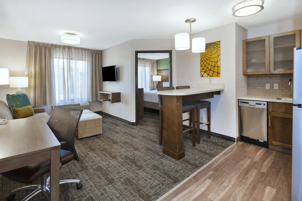 The image showcases a bright, modern living area with a kitchenette, a spacious bar with seating for dining or conversation, a dedicated work desk bathed in natural light, and thoughtfully chosen artwork that adds warmth and personality. The space is designed for both comfort and productivity, ideal for relaxing, enjoying a meal, or catching up on work.